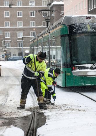 kuvassa kaksi miestä puhdistaa raitiotiekiskoja lumesta