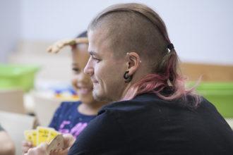Eftisledaren Oscar Maukkonen spelar kort med några barn. Han har kortsnaggat hår på sidorna, men på hjässan har han långt hår som samlats till en rosa hästsvans. I örat har en svart örring och ett piercingsmycke.