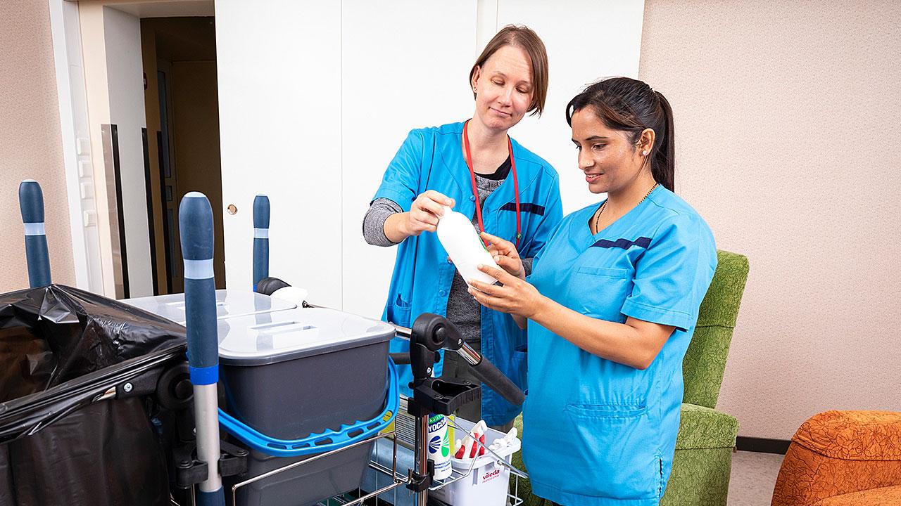 Palveluohjaaja Minna Felin ja opiskelija Shova Paudel katsovat pesuainepulloa työpaikallaan Kivelän sairaalassa