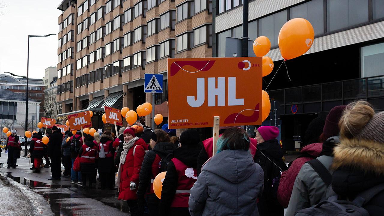 En lång kö med vinterklädda demonstranter trottoaren framför ett höghus. De bär orangea ballonger och en JHL-skylt.