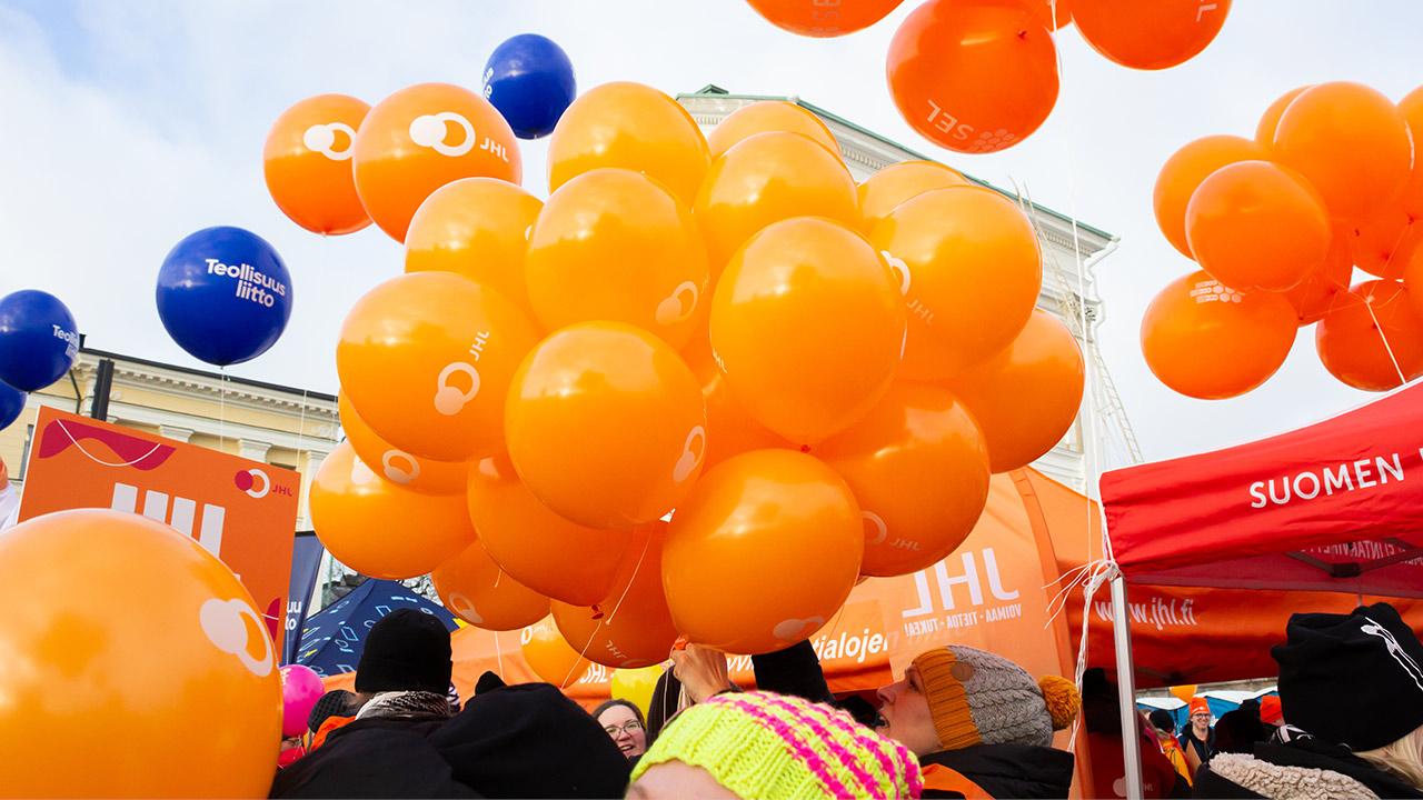 En klunga orangea ballonger flyger ovanför vinterklädda demonstranter.
