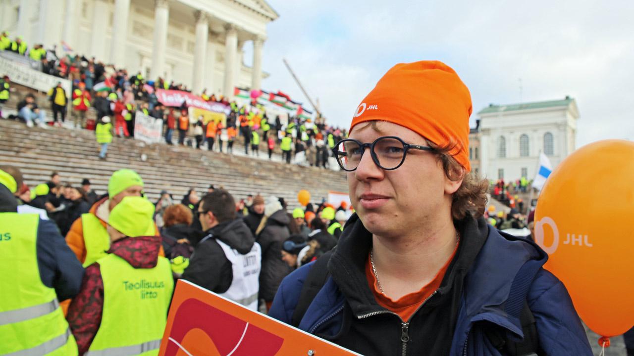 Person med orange mössa och glasögon vid domkyrkans trappa på Senatstorget. Trappan vimlar av demonstranter.