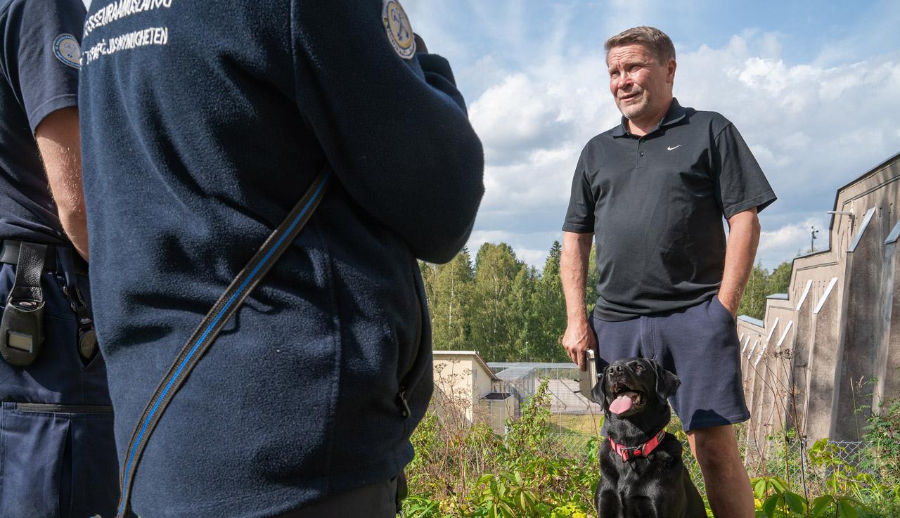 Antti Santamäki katsoo koira vierellään selin kameraan seisovia vankilavirkailijoita.