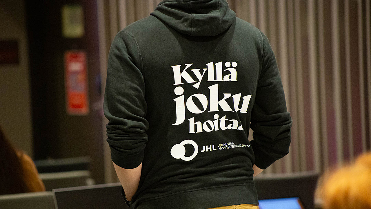 Henkilö seisoo selin yllään ammattiliitto JHL:n huppari, jossa lukee Kyllä joku hoitaa.