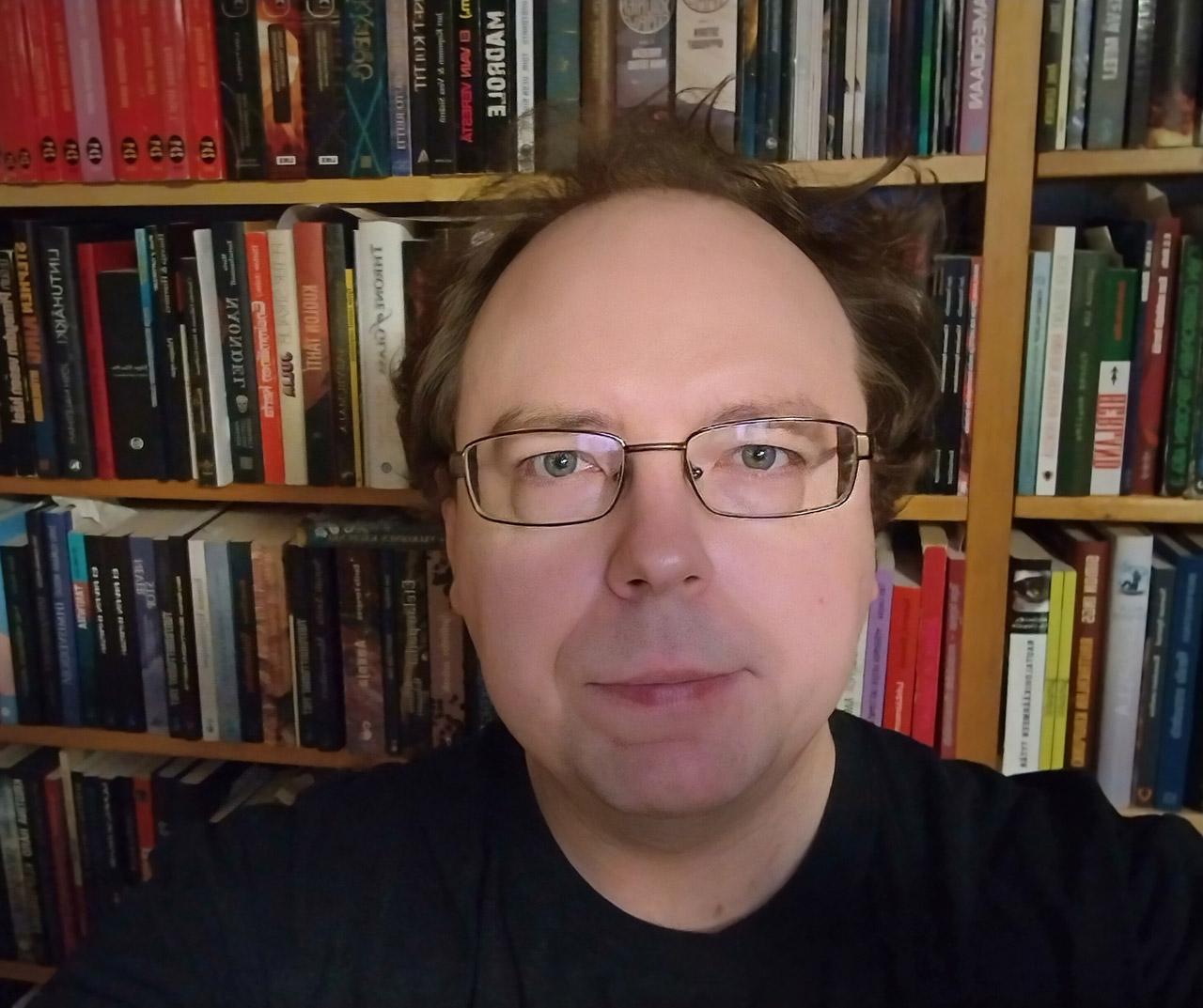 Kirjastotyöntekijä Matti Järvinen katsoo kameraan taustallaan kirjahylly.