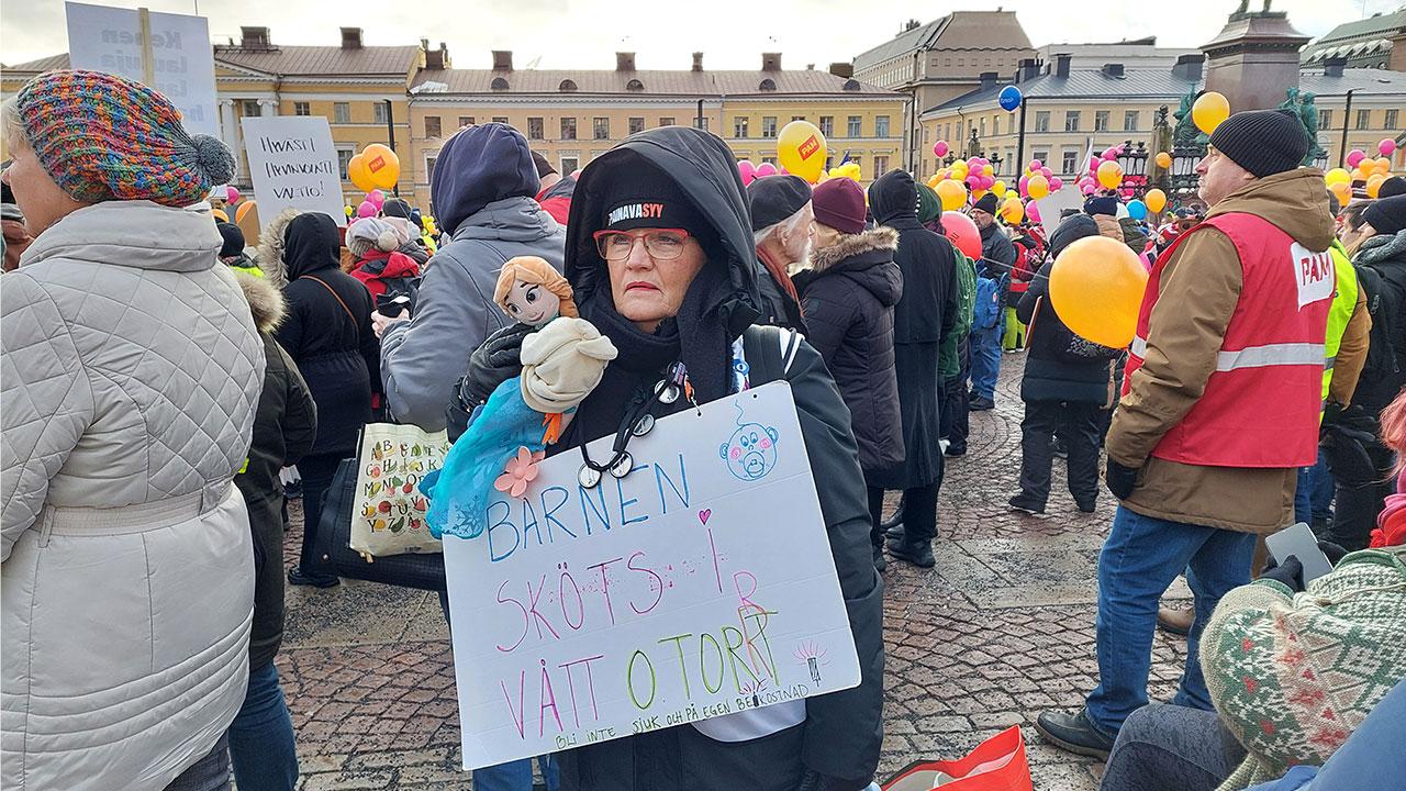 Vinterklädd demonstrant på torget håller en skylt med texten "Barnen sköts i vått och torrt".