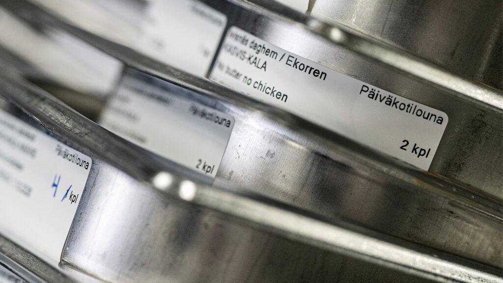 Teräksisten astioiden kylkeen on teipattu sen koulun tai päiväkodin nimi, minne ruoka on matkalla.
