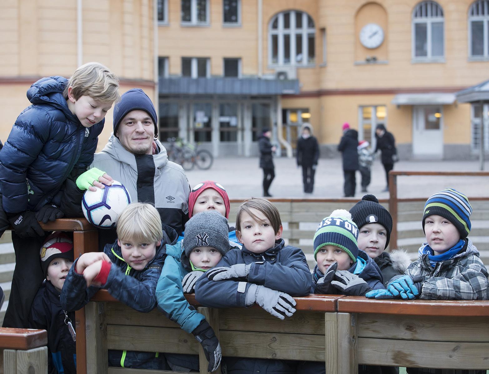 Iltapäiväkerhon ohjaaja Oscar Maukkonen poseeraa pienessä jalkapallokaukalossa lasten kanssa.