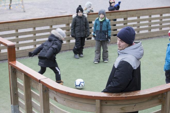 Iltapäiväkerhonohjaaja Oscar Maukkonen pelaa lasten kanssa jalkapalloa pienessä jalkapallokaukalossa.