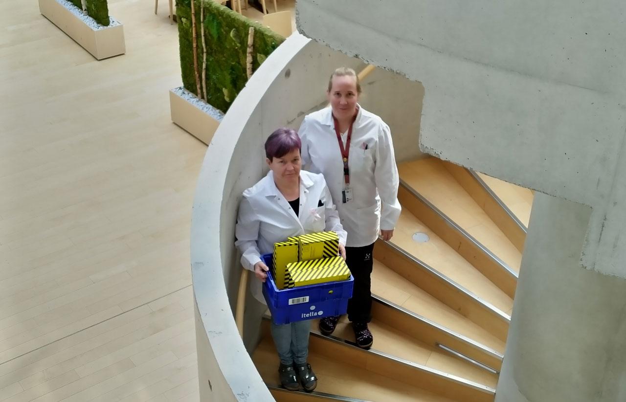 JHL-anslutna laboratoriearbetarna Merja Hautala och Tiina Peltonen står i en spiraltrappa på Livsmedelsverket med material anslutande till coronavirustester i en låda. 