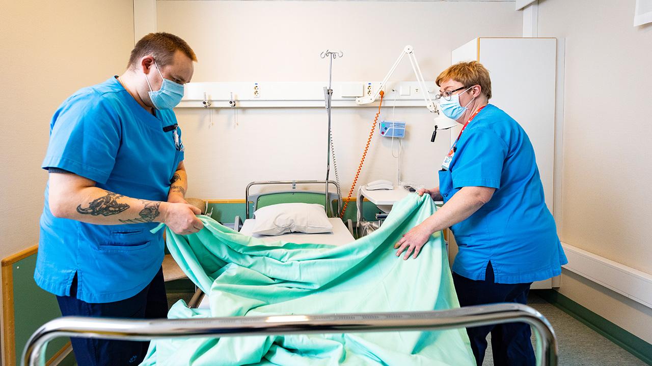 Mikael Schön och Ann-Christin Wikholm bäddar en sjukhussäng. De är klädda i blå, kortärmade tröjor ocjh svarta byxor. Sängen har grönt överkast. 