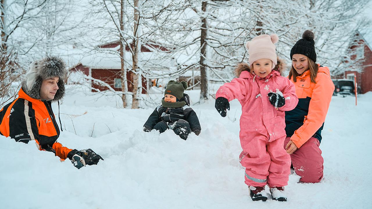 Isä.äiti ja kaksi pientä lasta leikkivät lumileikkejä Posiolla.  Taustalla punaisia puurakennuksia. 